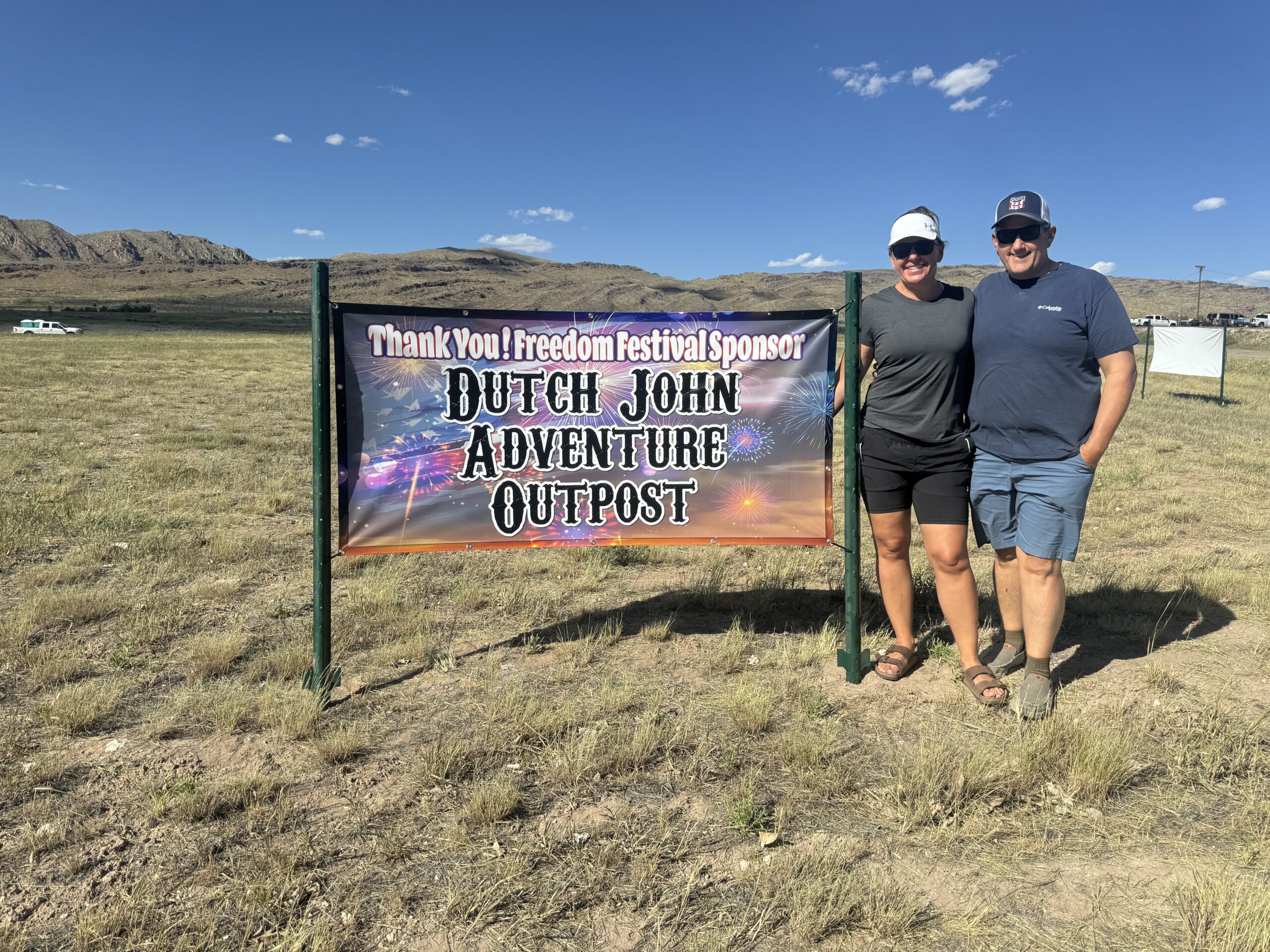 Andy and Kelly Morse, owners of Dutch John Adventure Outpost, standing proudly beside a sponsorship sign at an outdoor event, with the scenic mountains of Dutch John, Utah in the background.